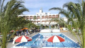 A pool area with lounge chairs and umbrellas, surrounded by palm trees and a building with arches in the background. - Hotel Village Club Santa Caterina (Scalea)