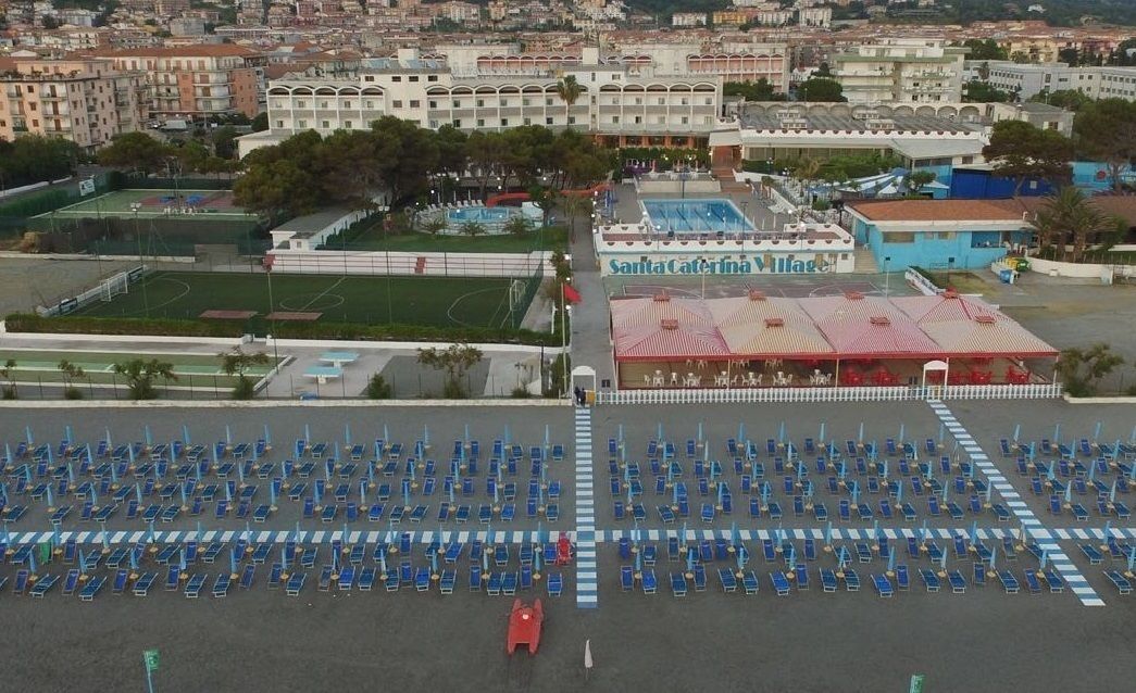 Un amplio espacio exterior con numerosas sillas azules dispuestas en filas, un campo deportivo y un edificio con tejado rojo identificado como “Santa Caterina Village”.