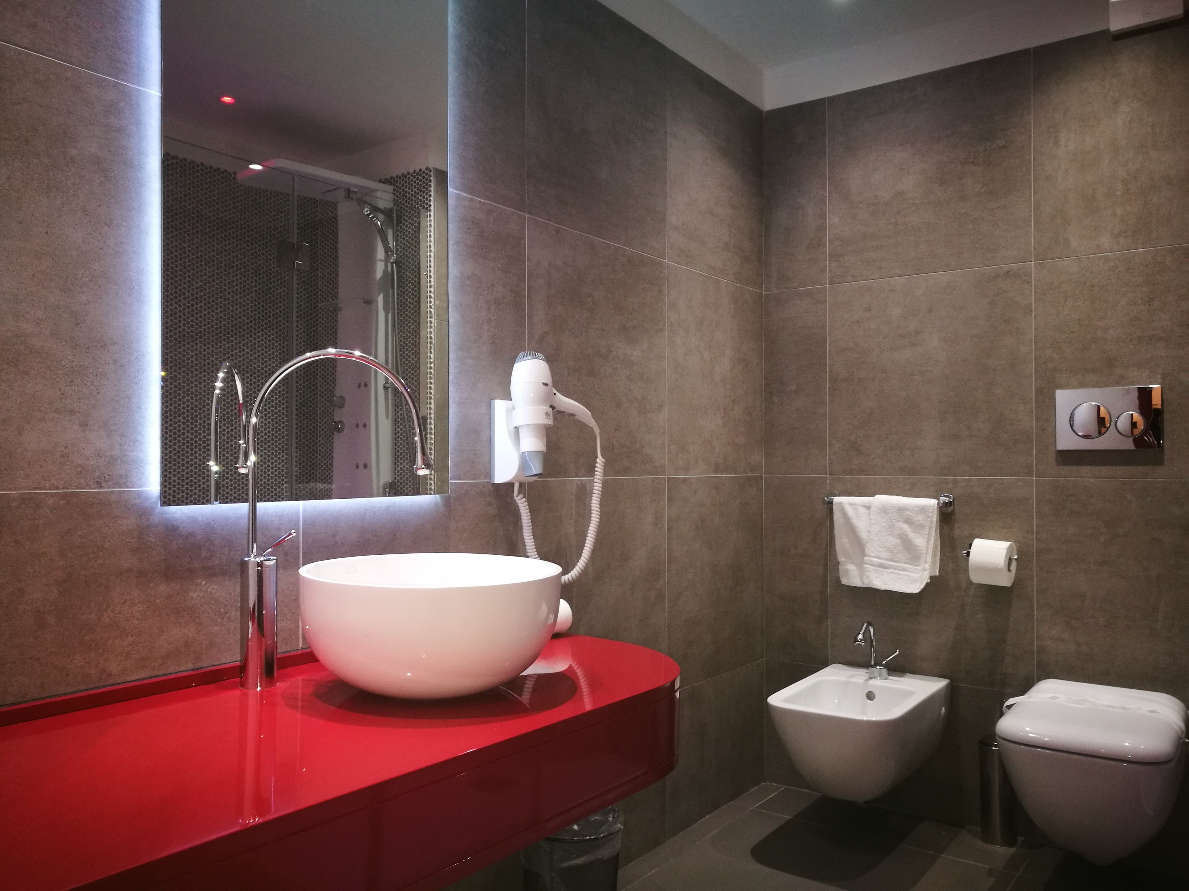 Ein modernes Badezimmer mit einem großen Spiegel, einer roten Ablagefläche, einem weißen Waschbecken, einem Bidet und einer Toilette.