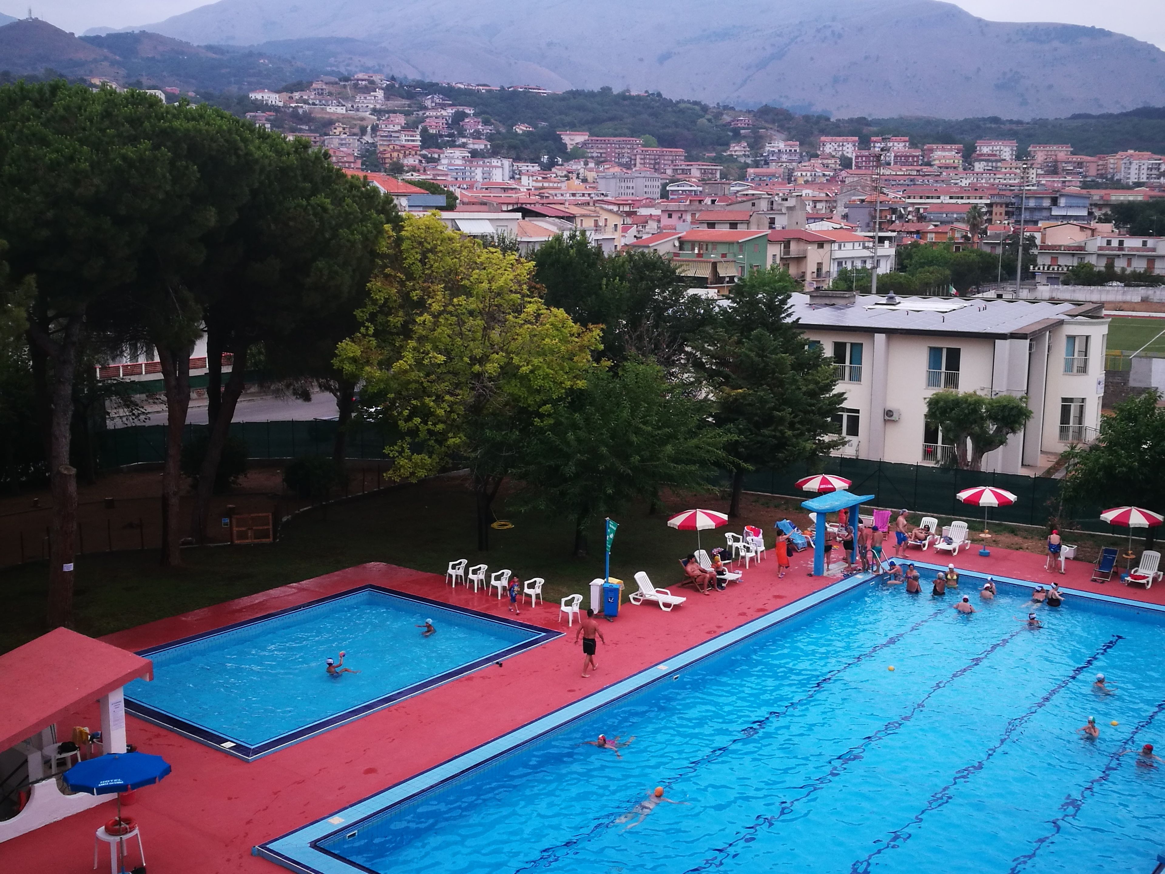 A swimming pool with multiple lanes, surrounded by lounge chairs and umbrellas, set against a backdrop of a residential area with buildings and trees.