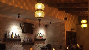 A bar area with a brick wall, wooden ceiling, and hanging lamps. There are shelves with bottles and glasses, and wicker chairs.