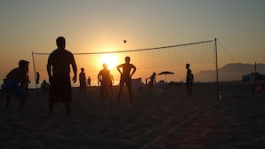 A beach volleyball game at sunset with players silhouetted against the sun.