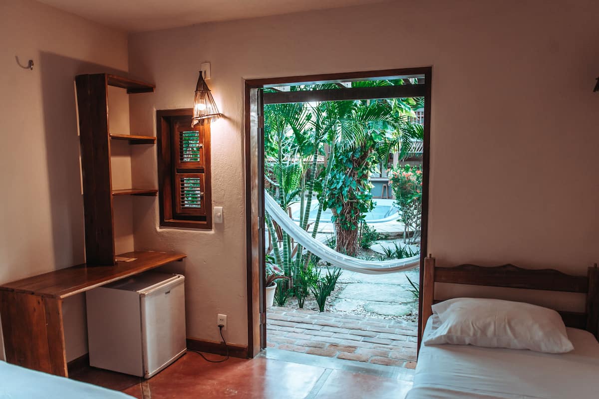 Quarto com cama, escrivaninha pequena, frigorífico, prateleira na parede e porta que dá para um jardim com rede.