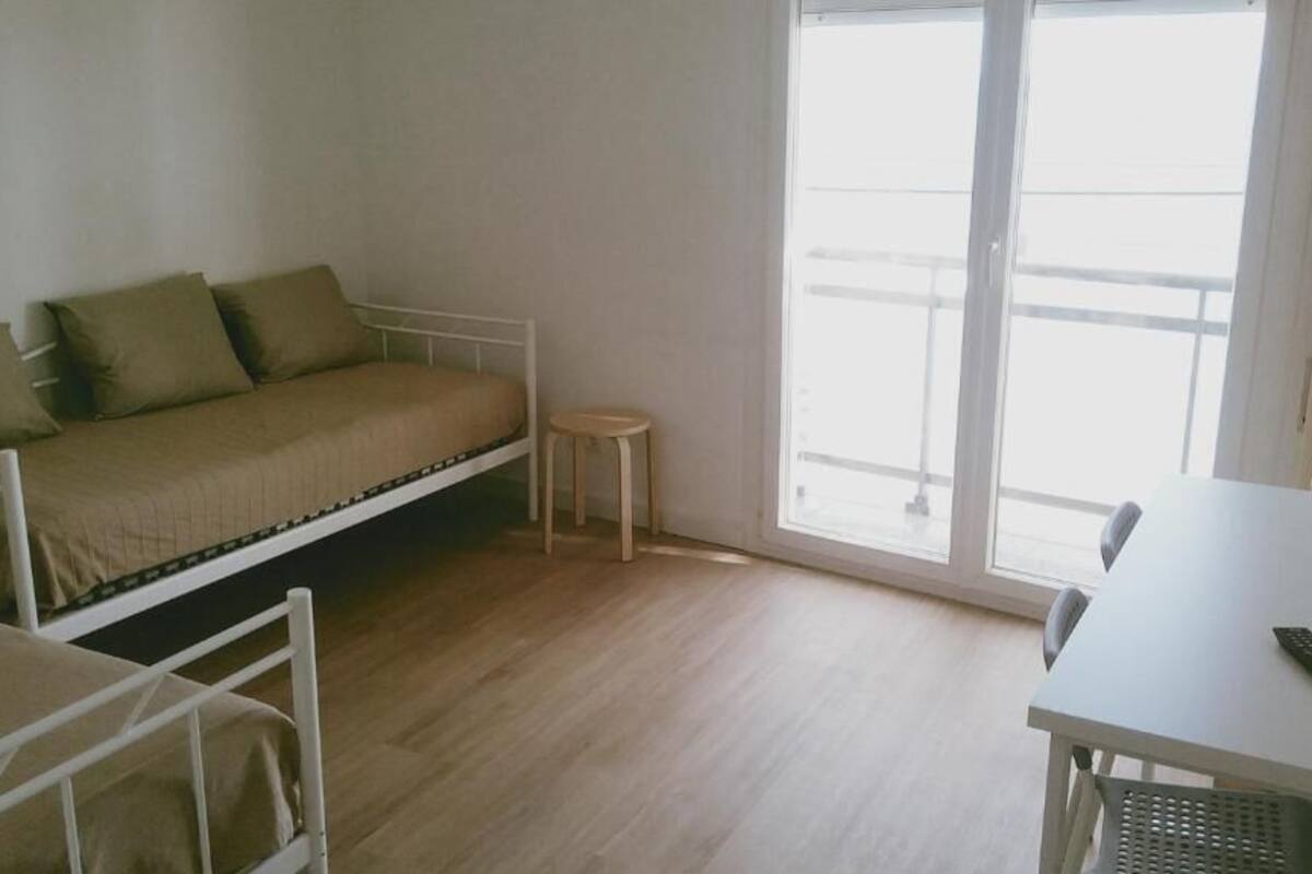 A room with a bed, a small stool, a table, and a window with a view.