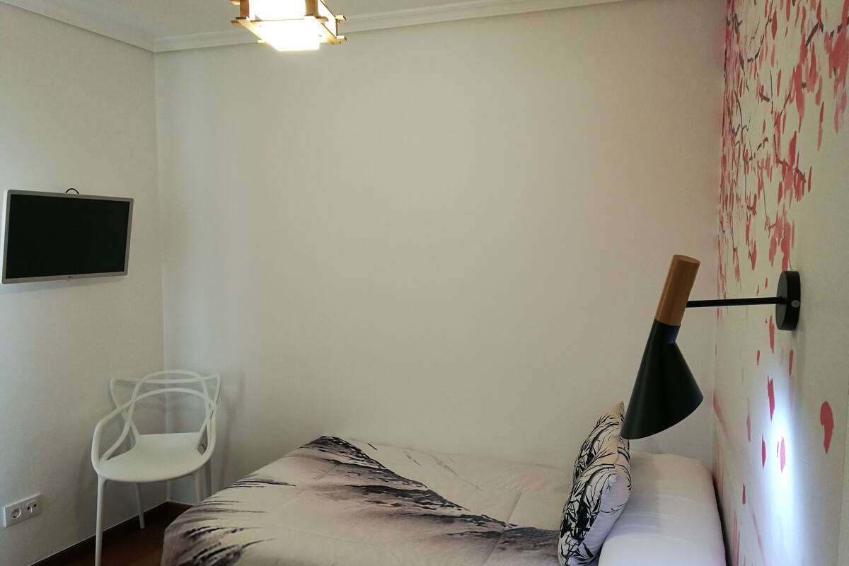 A bedroom with a bed, a chair, a TV, and a wall-mounted lamp.