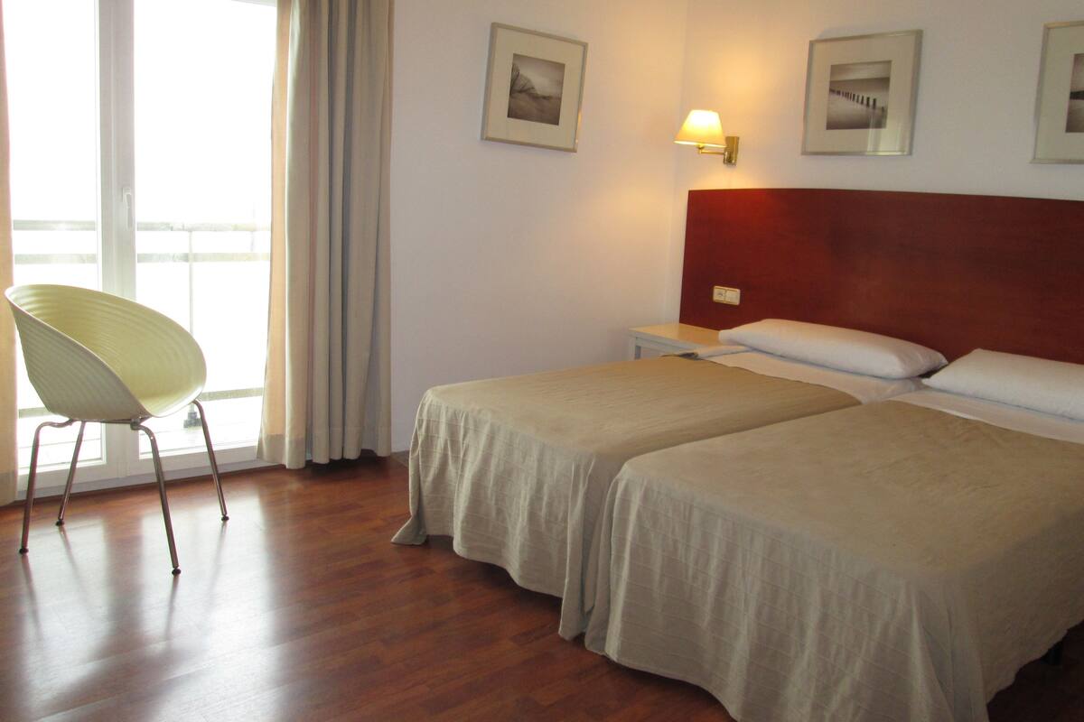 A hotel room with two beds, a chair, a nightstand, and a window with curtains.
