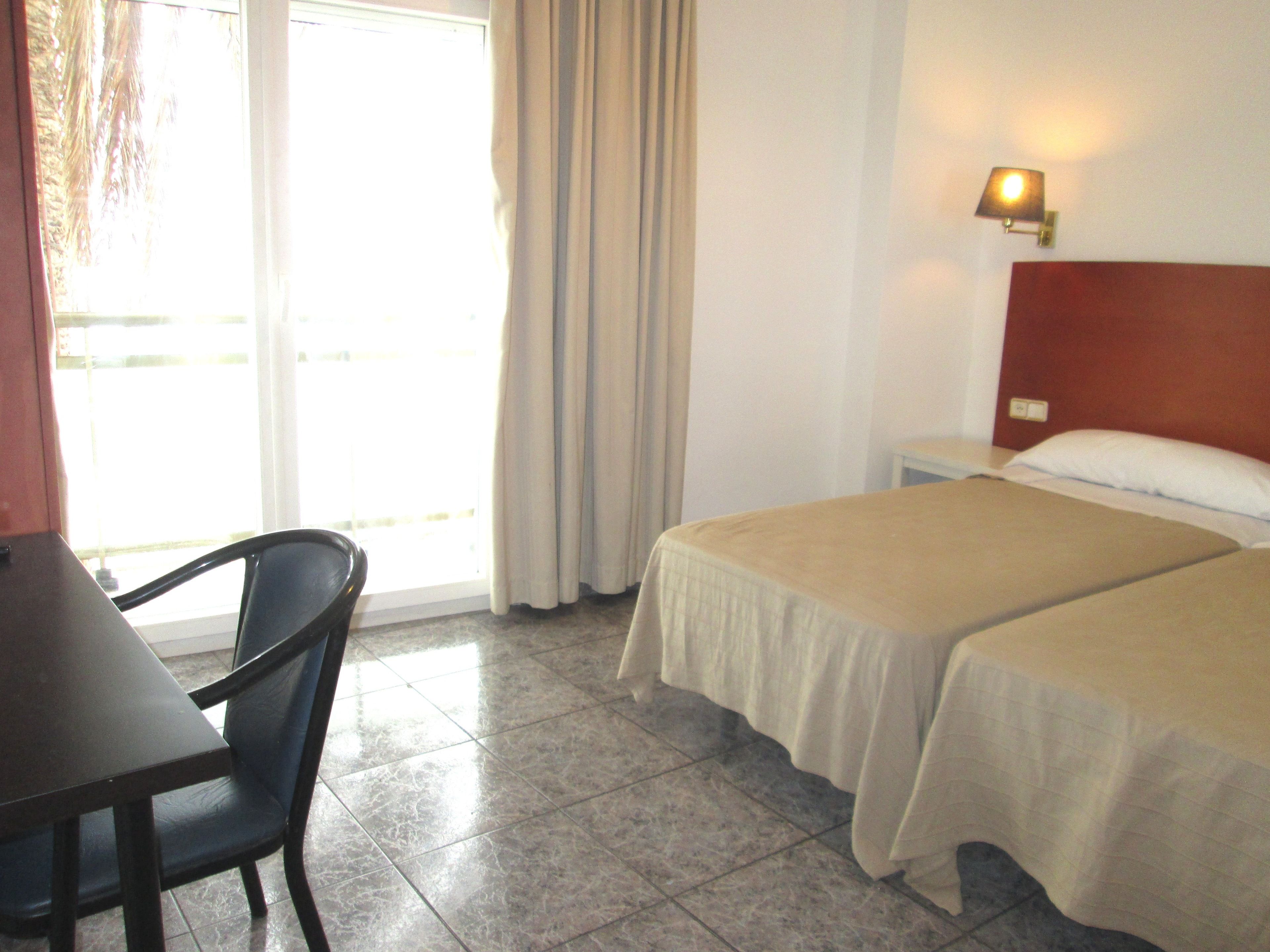 Habitación de hotel con dos camas, un escritorio, una silla, un ventanal con cortinas y una lámpara de pared.