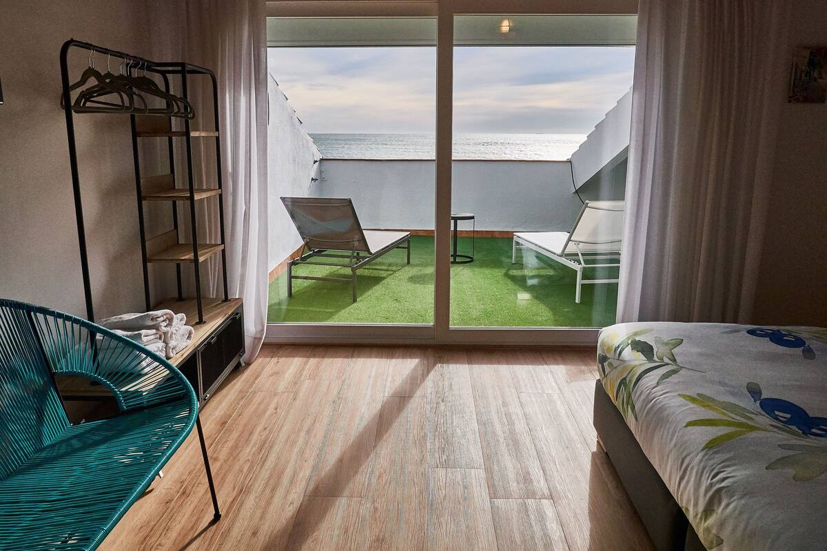 A room with a bed, a green chair, a small table, a balcony with two chairs, and a metal rack with hangers.