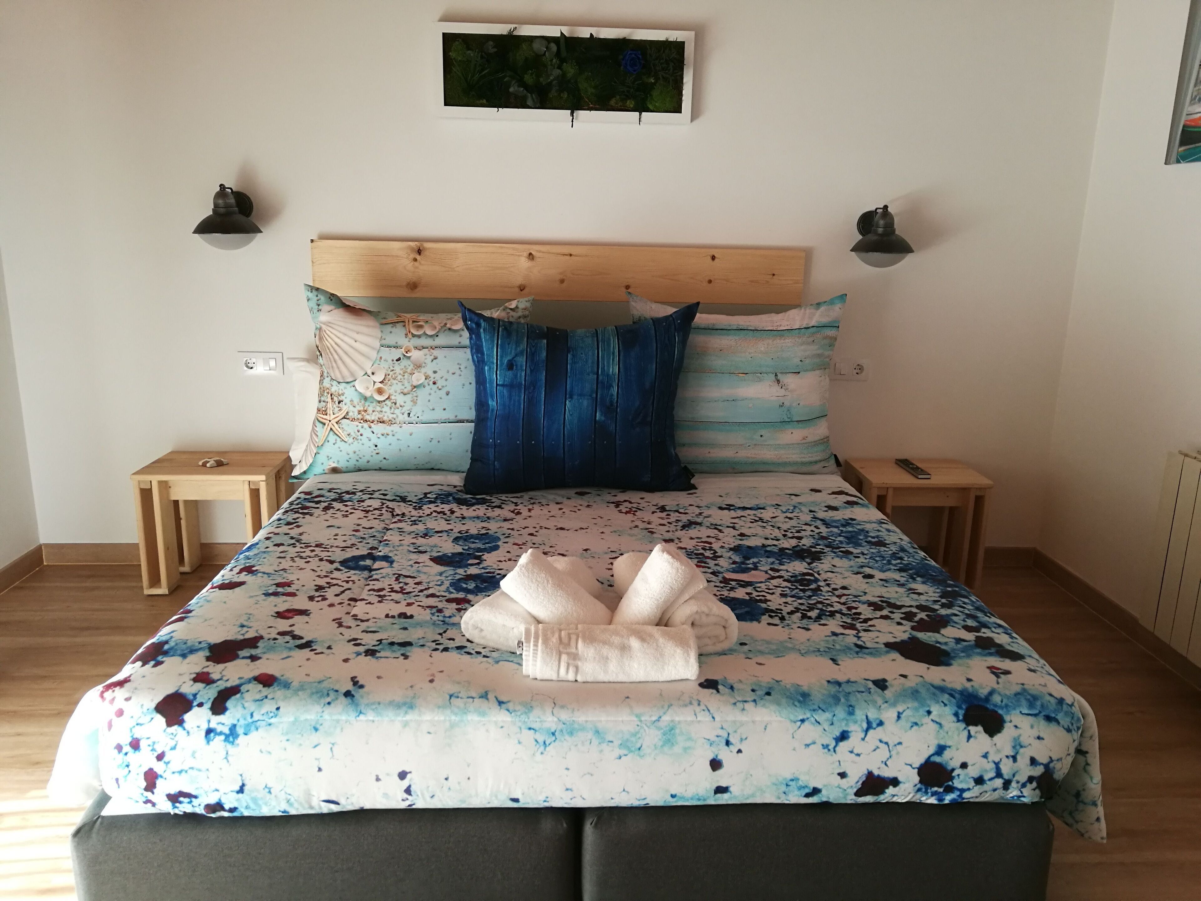 Un dormitorio con cabecera de madera, una cama con colcha azul y blanca, dos mesitas de noche y un cuadro colgado en la pared.