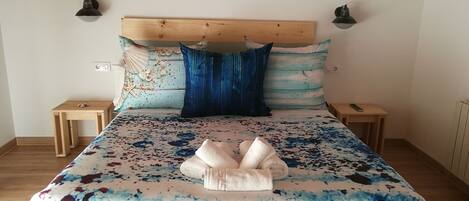 Un dormitorio con cabecera de madera, una cama con colcha azul y blanca, dos mesitas de noche y un cuadro colgado en la pared.