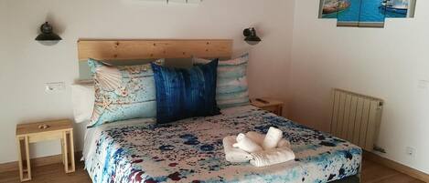 A bedroom with a wooden headboard, a bed with a floral bedspread, two wall-mounted lamps, a small bedside table, and a painting of a harbor on the wall.