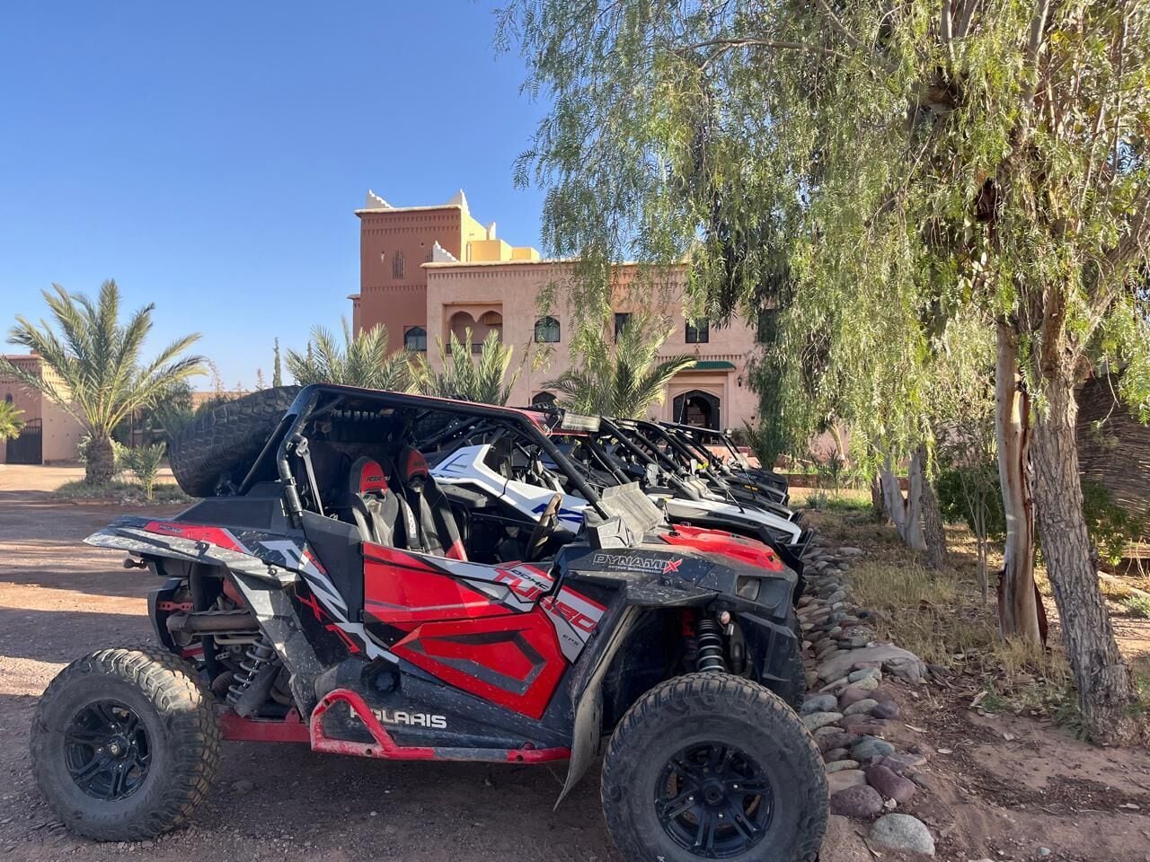 Um veículo off-road Polaris RZR vermelho e preto estacionado em ambiente natural, com um edifício ao fundo.