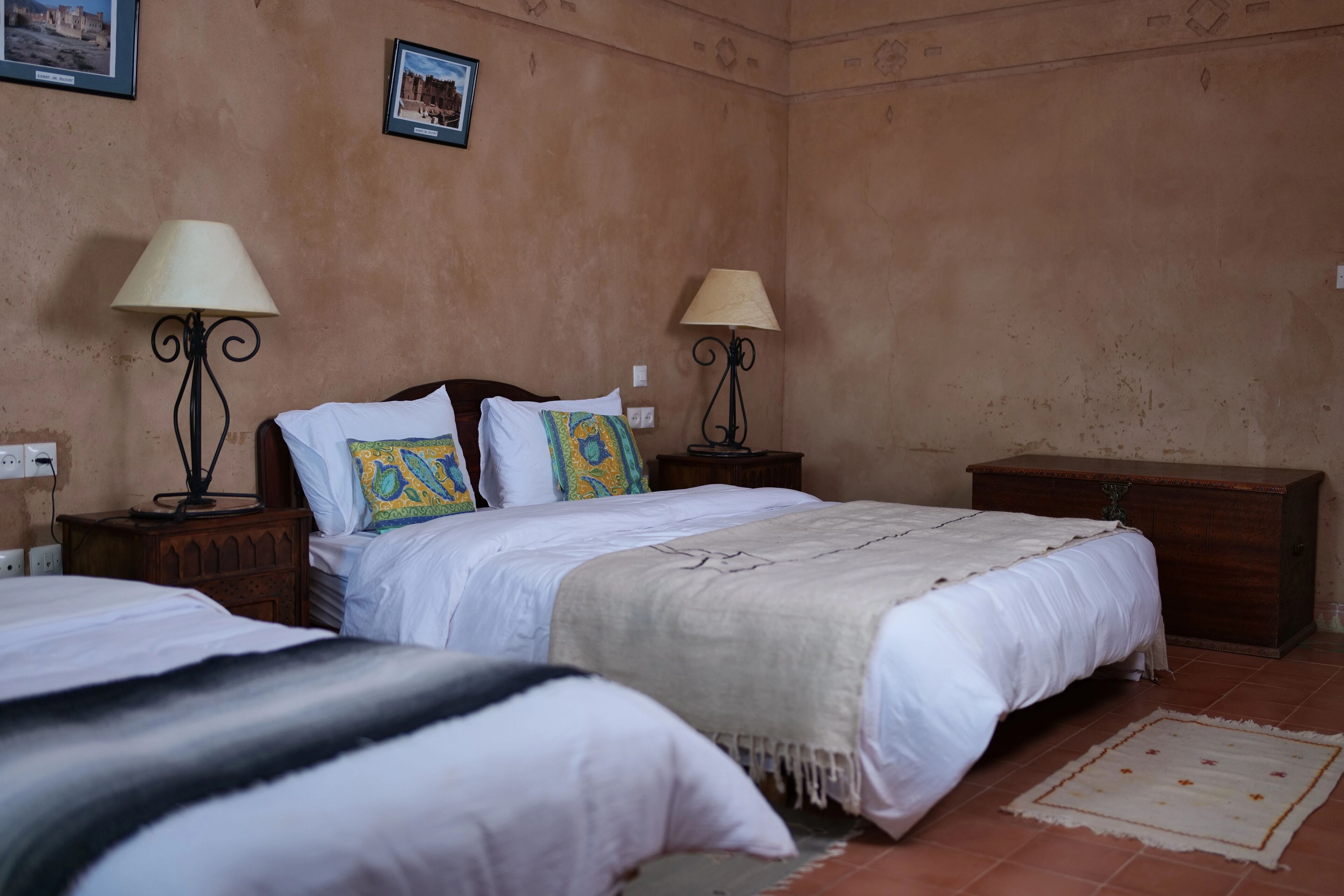 A bedroom with two beds, each with a bedside table and lamp, a wooden dresser, and a tiled floor.