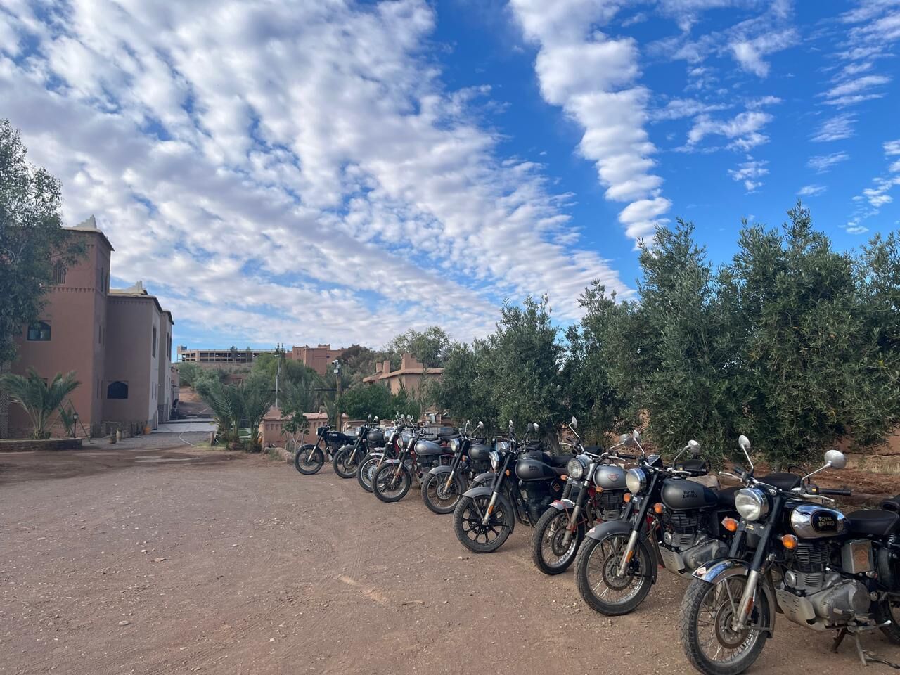 Una fila de motocicletas estacionadas en un camino de tierra, con un edificio y árboles al fondo.