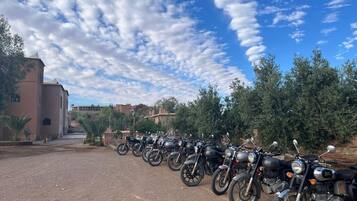 Una fila de motocicletas estacionadas en un camino de tierra, con un edificio y árboles al fondo.