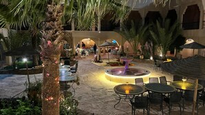 Un área de patio al aire libre con mesas y sillas, una fuente central y palmeras.