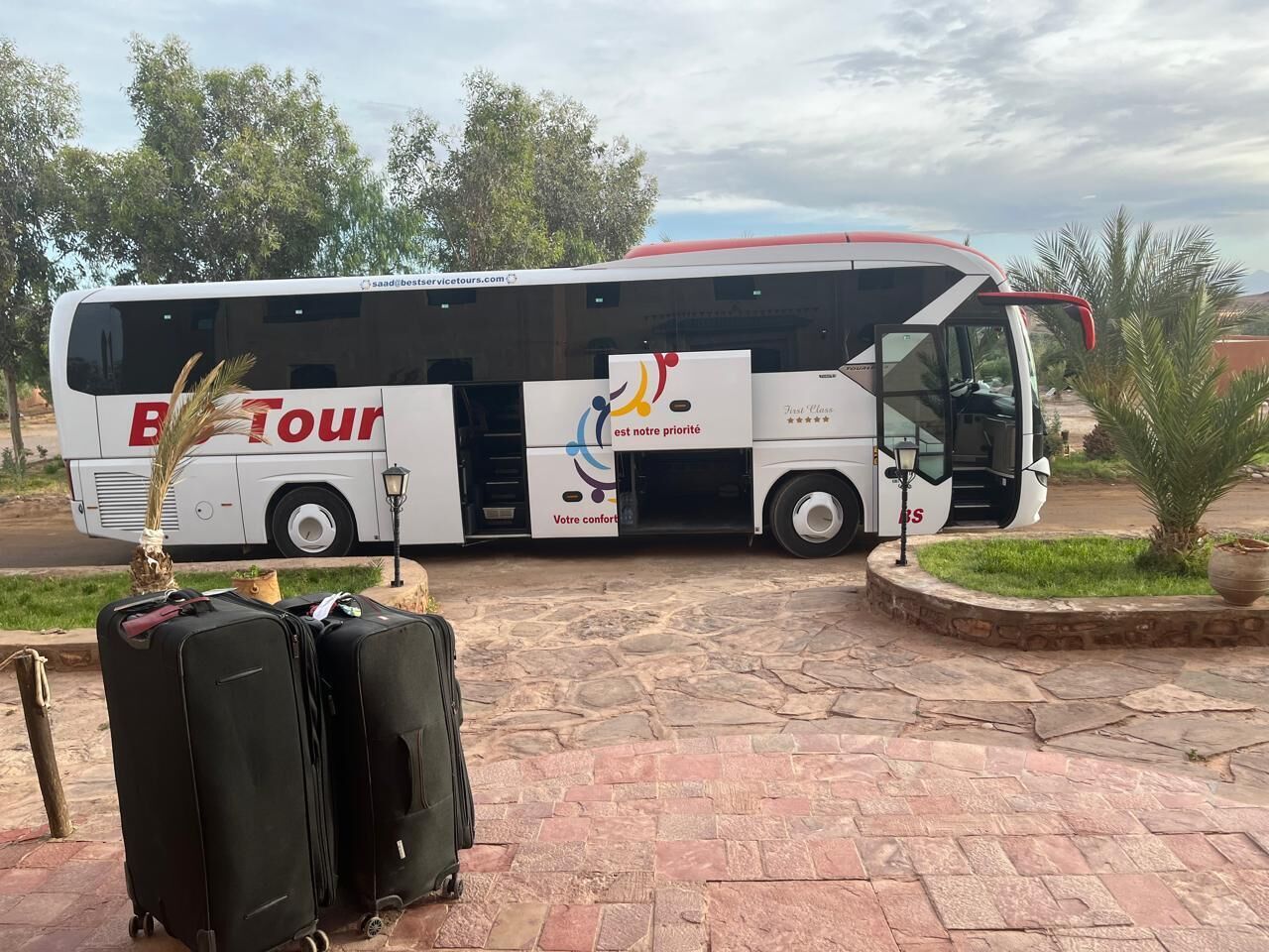 A tour bus with luggage in front, parked on a paved area with trees and a building in the background.