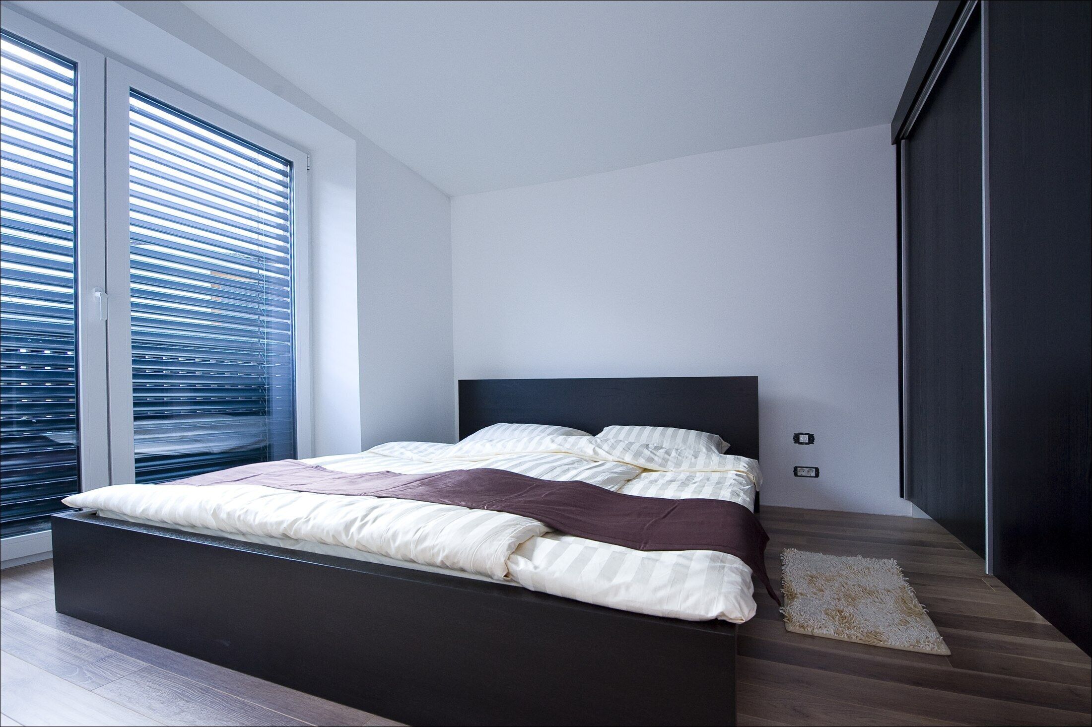 A modern bedroom with a large bed, a dark headboard, and a view through floor-to-ceiling windows.