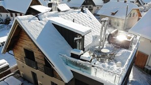 Un bâtiment moderne doté d’un toit en pente recouvert de neige, d’une terrasse fermée par des baies vitrées et aménagée avec du mobilier d’extérieur, et d’une cheminée bien visible.