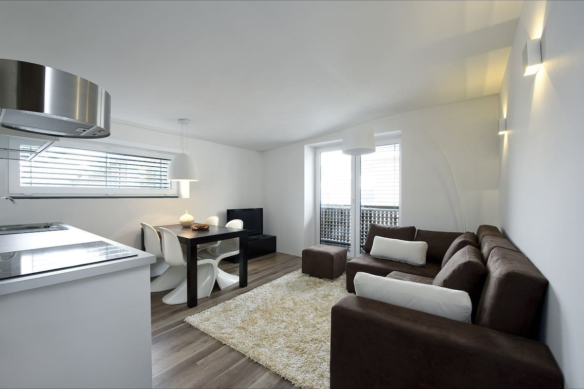 Un salon moderne avec un canapé marron, un coin repas avec une table et des chaises, et une cuisine équipée d’une cuisinière et d’un téléviseur.