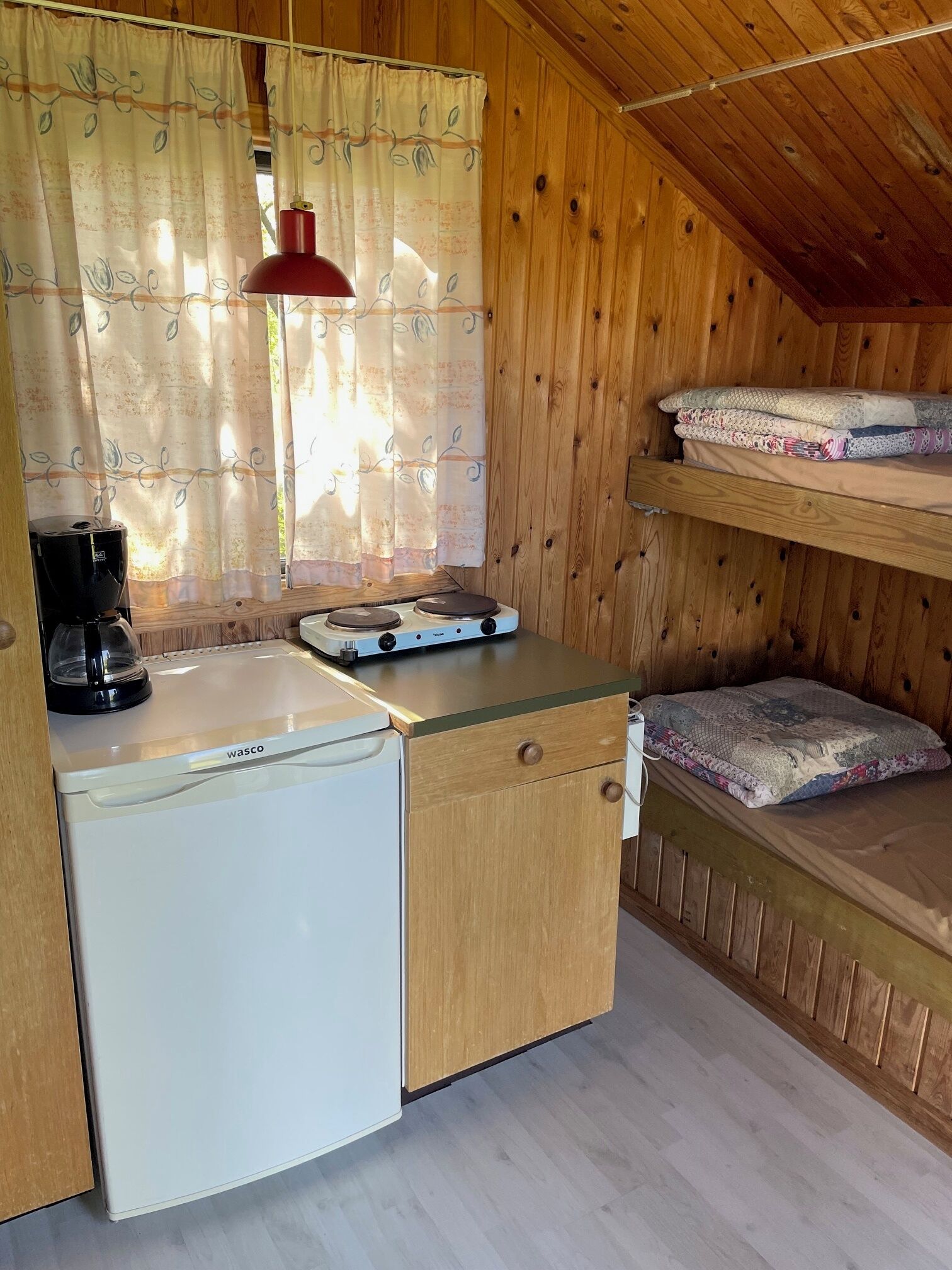 Una kitchenette compacta con un refrigerador blanco, una cafetera y una estufa pequeña. Hay un gabinete de madera con un cajón y un estante arriba. La habitación tiene paredes de madera y un techo inclinado.