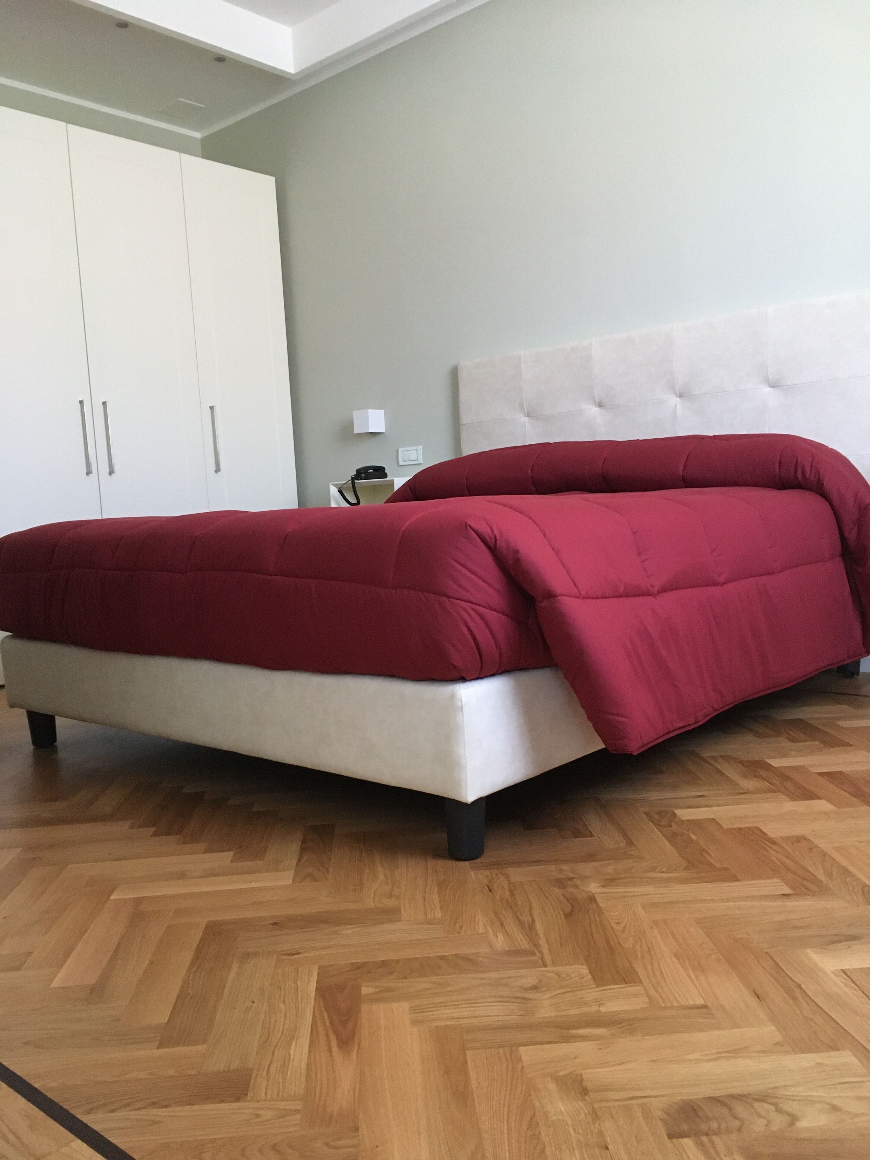 Un lit avec une couverture rouge, une tête de lit blanche et un parquet en bois disposé en motif chevrons.