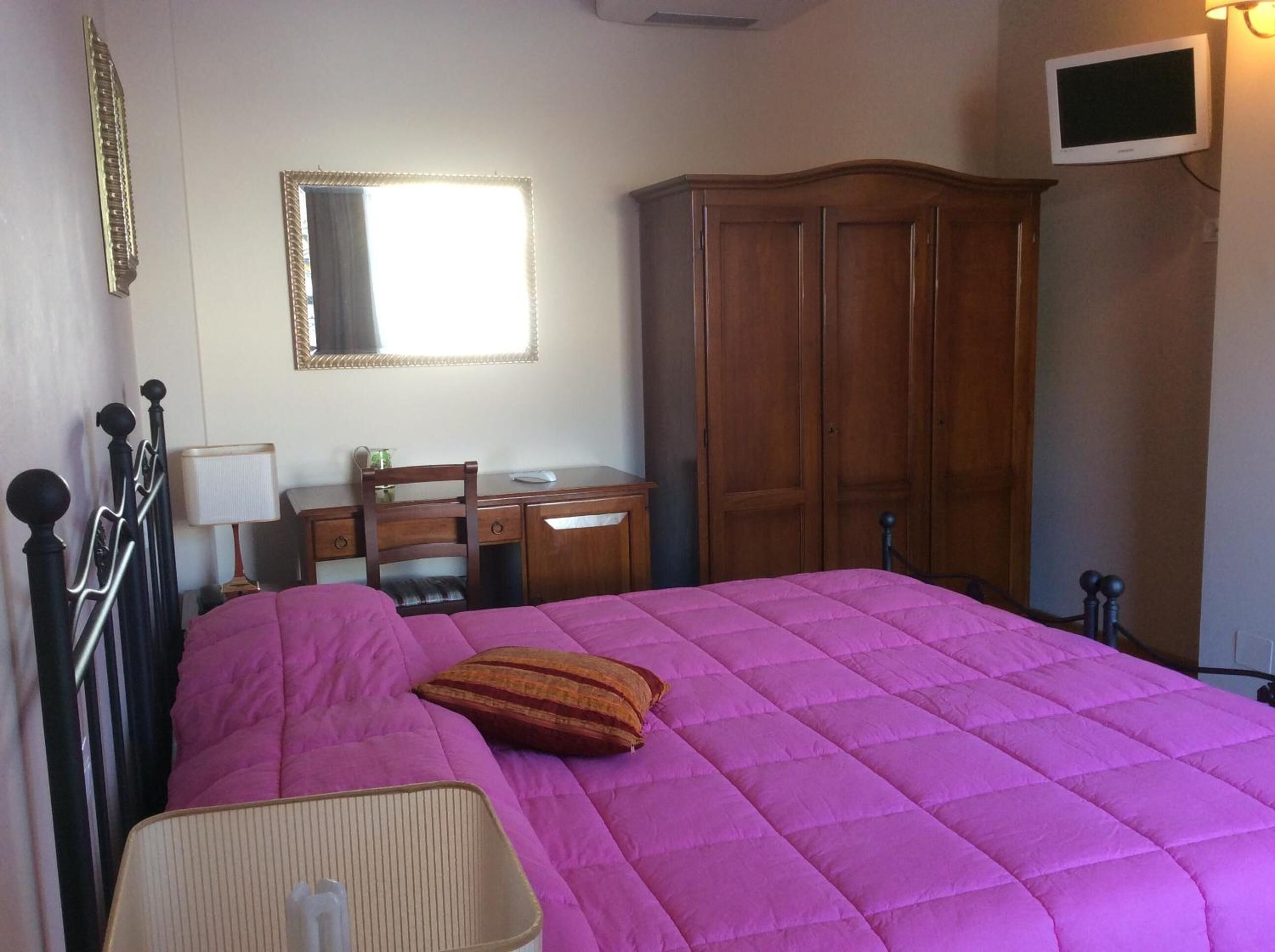 Un dormitorio con una cama grande, un armario de madera, un escritorio con una silla, un espejo y un televisor en la pared.