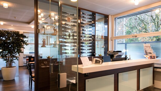 Un espace d’accueil avec une cloison en verre, un comptoir de réception, une plante en pot et une vue sur les arbres à l’extérieur.