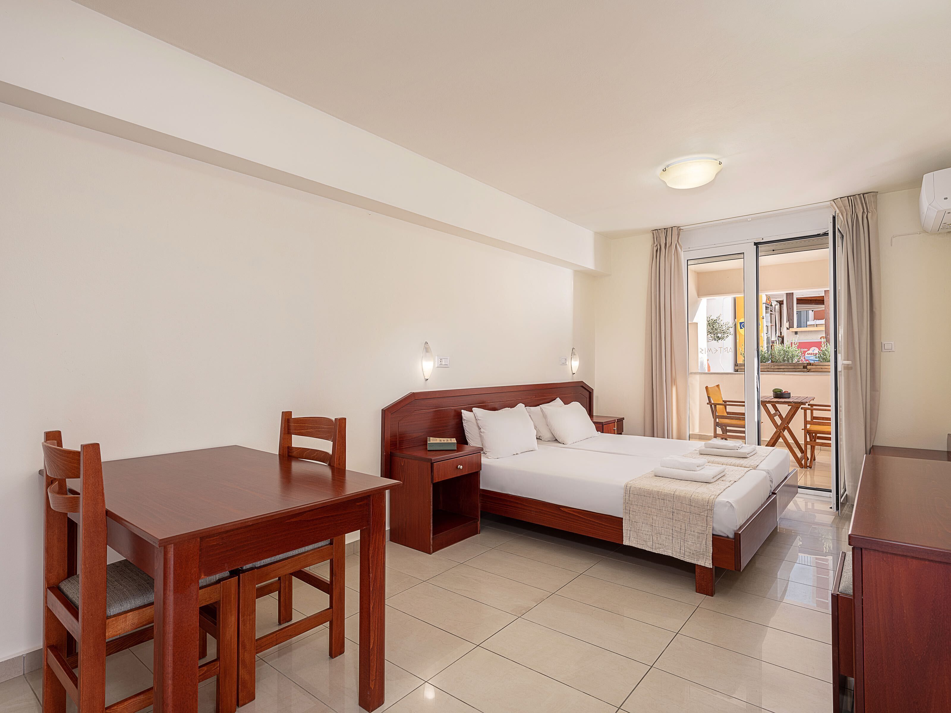 A hotel room with a wooden bed, a wooden desk, and a chair. There is a window with curtains, a door leading to a balcony, and a ceiling light.