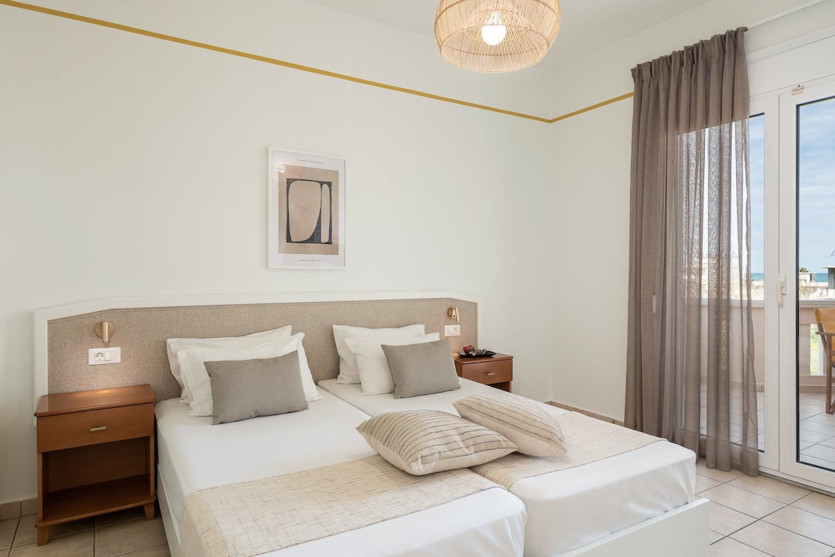 A hotel room with a large bed, bedside tables, a nightstand, a ceiling light fixture, and a view of the outdoors through a sliding glass door.