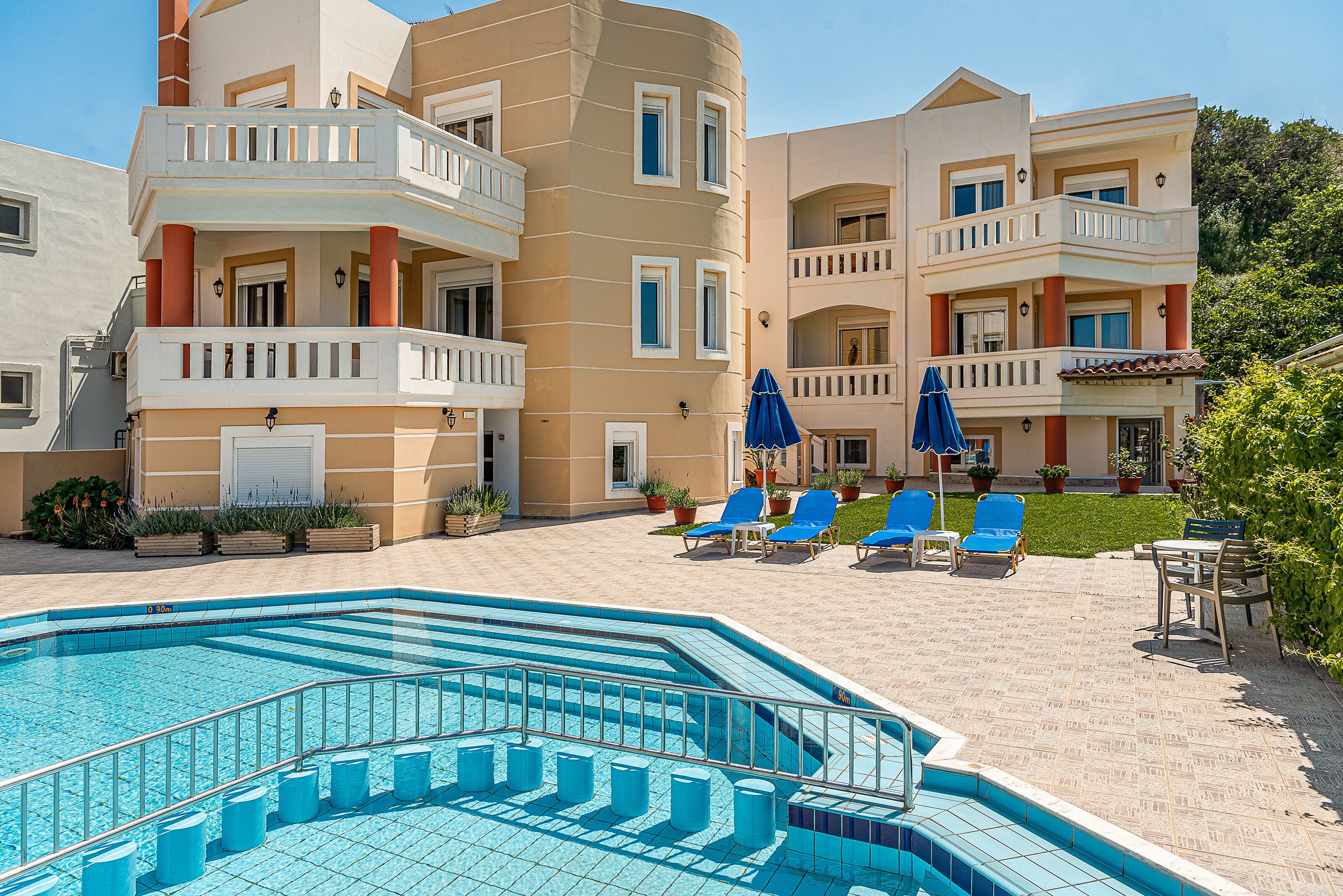 A swimming pool with blue tiles, surrounded by lounge chairs and umbrellas, and a building with balconies and windows.