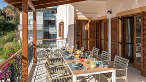 A terrace with a wooden table set for dining, surrounded by chairs, with a view of a building and greenery in the background. - Artemis Apartments (Chania)