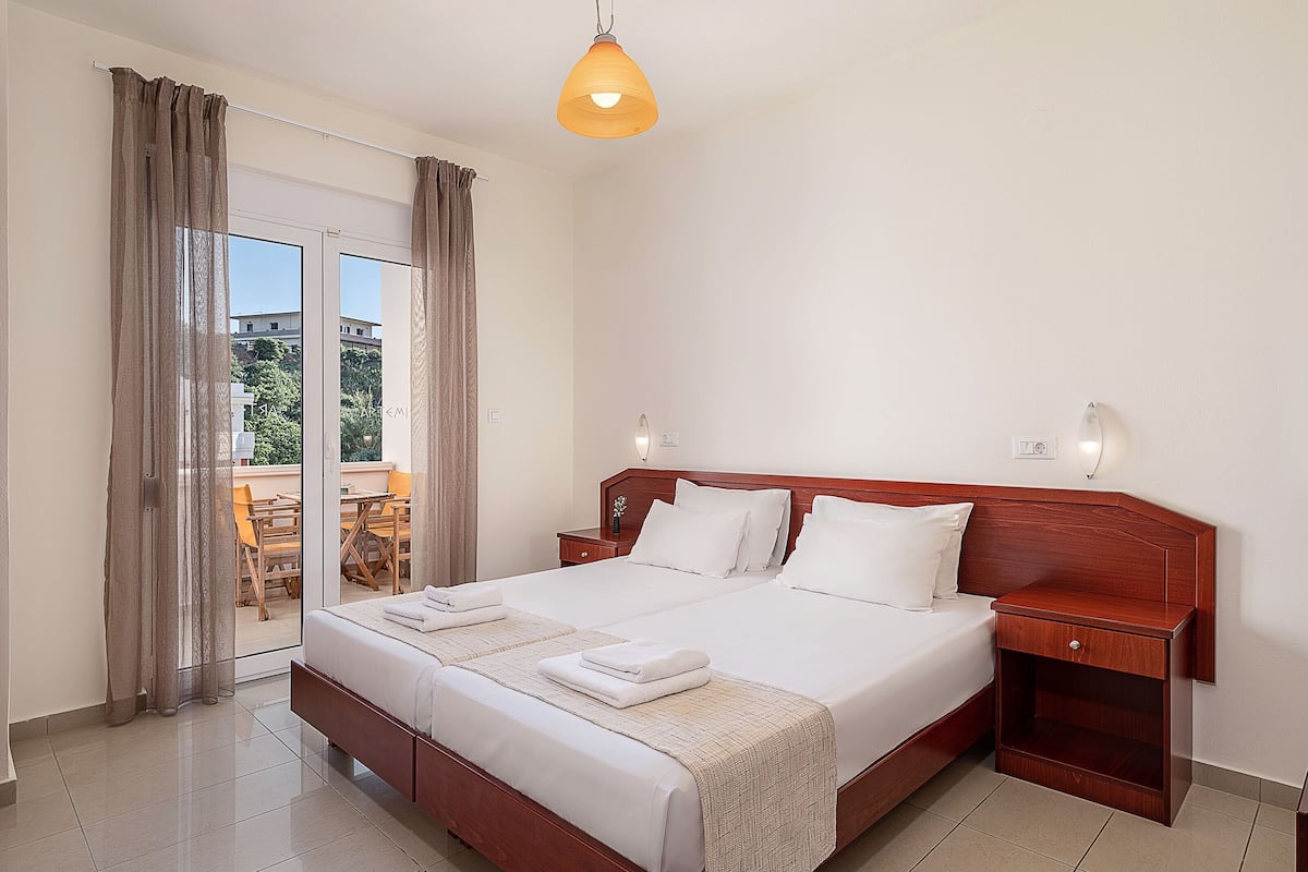 A hotel room with a large bed, wooden headboard, two bedside tables, a nightstand, and a balcony with a table and chairs.