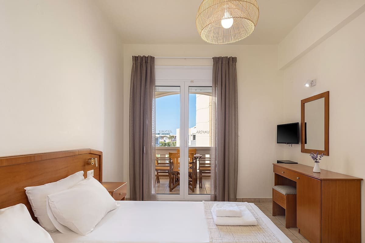 A hotel room with a large bed, a wooden nightstand, a TV, a mirror, and a view of the outside through a glass door.