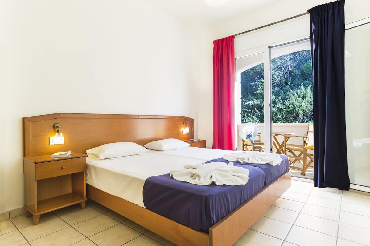 A hotel room with a wooden bed, a nightstand, a bedside lamp, a window with red curtains, and a view of greenery outside.