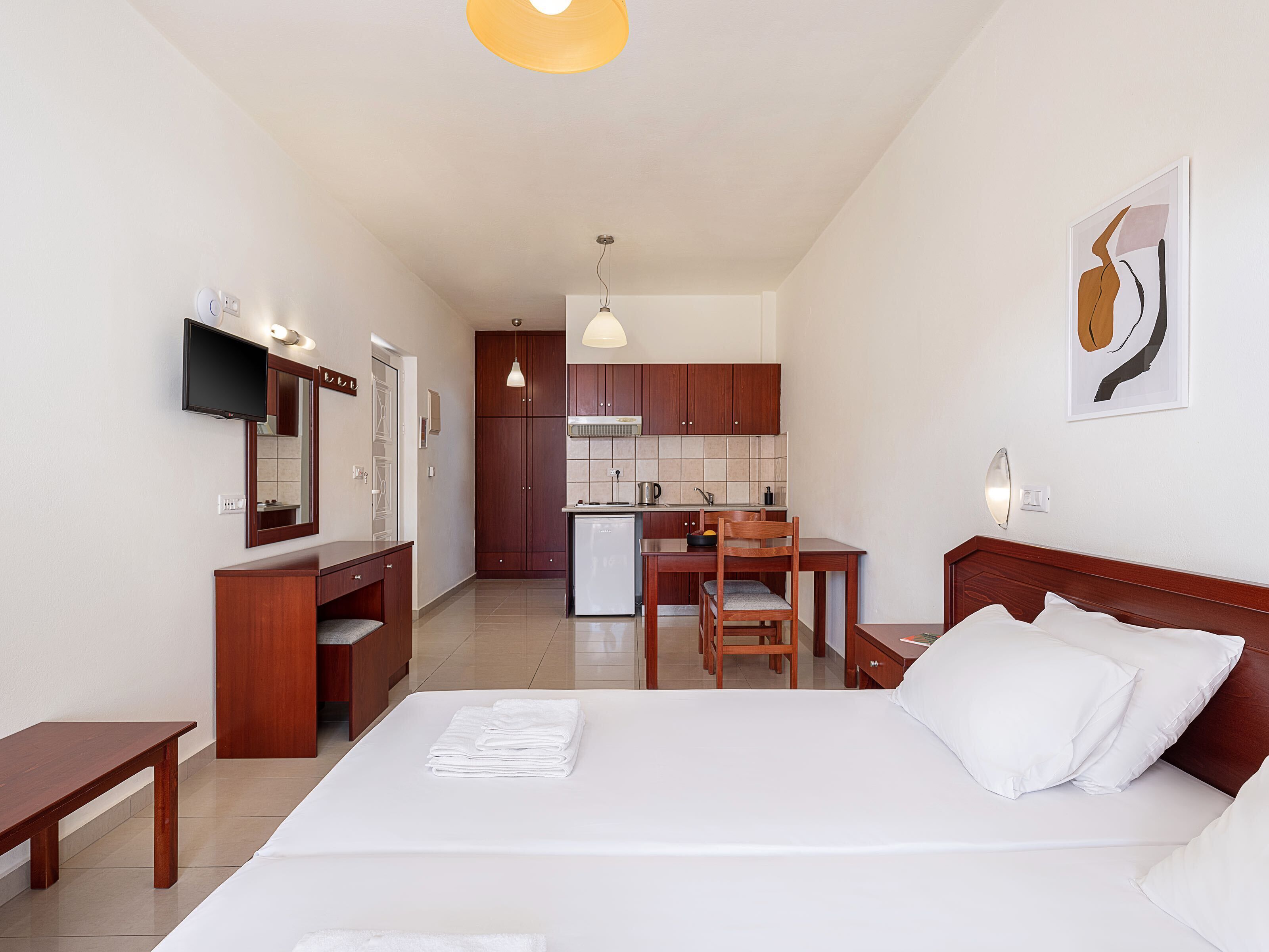 A hotel room with a bed, a desk, a chair, a small kitchenette, and a TV.