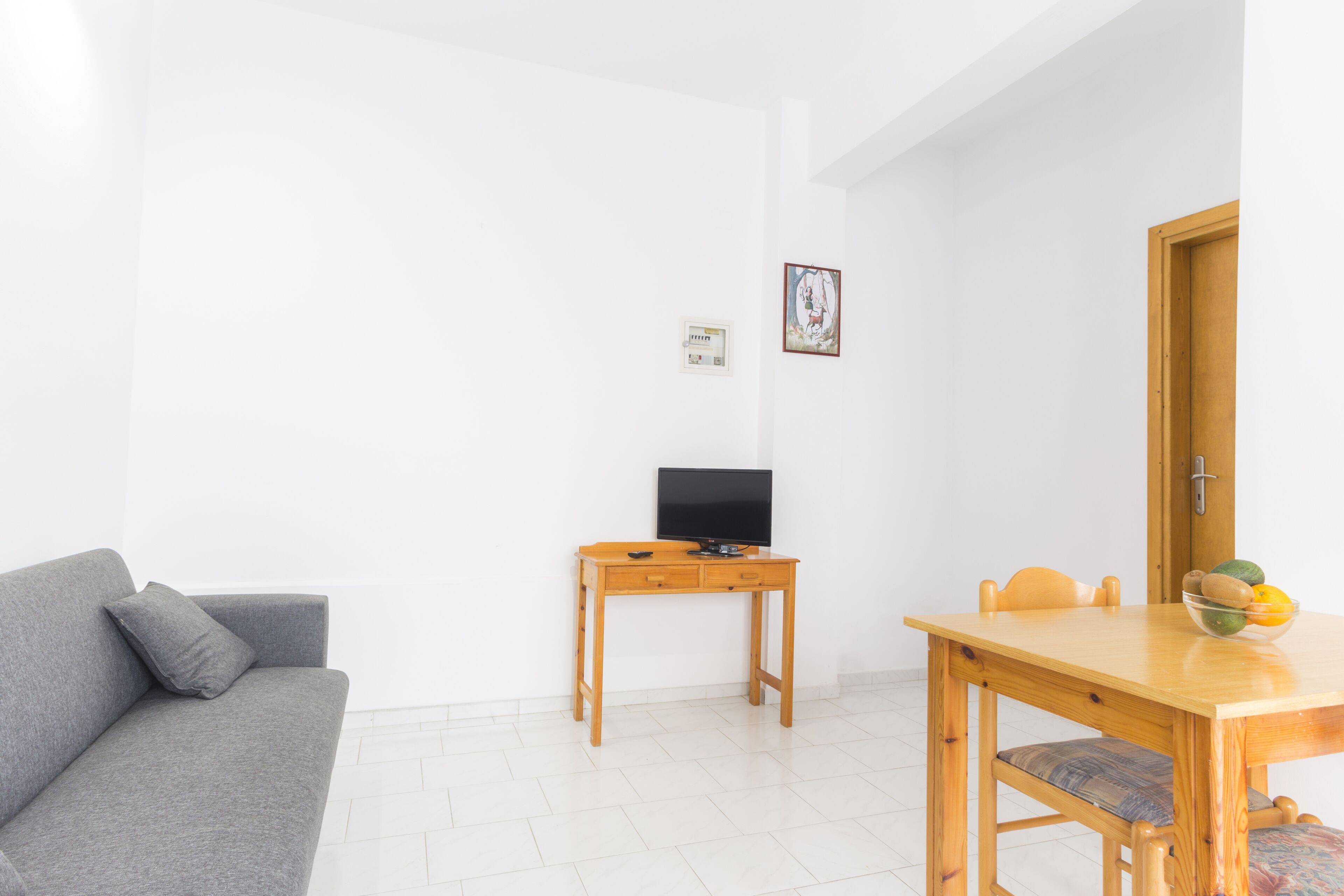 A minimalist living room with a grey sofa, a wooden TV stand with a flat-screen TV, a wooden dining table with chairs, and a small fruit bowl on the table.