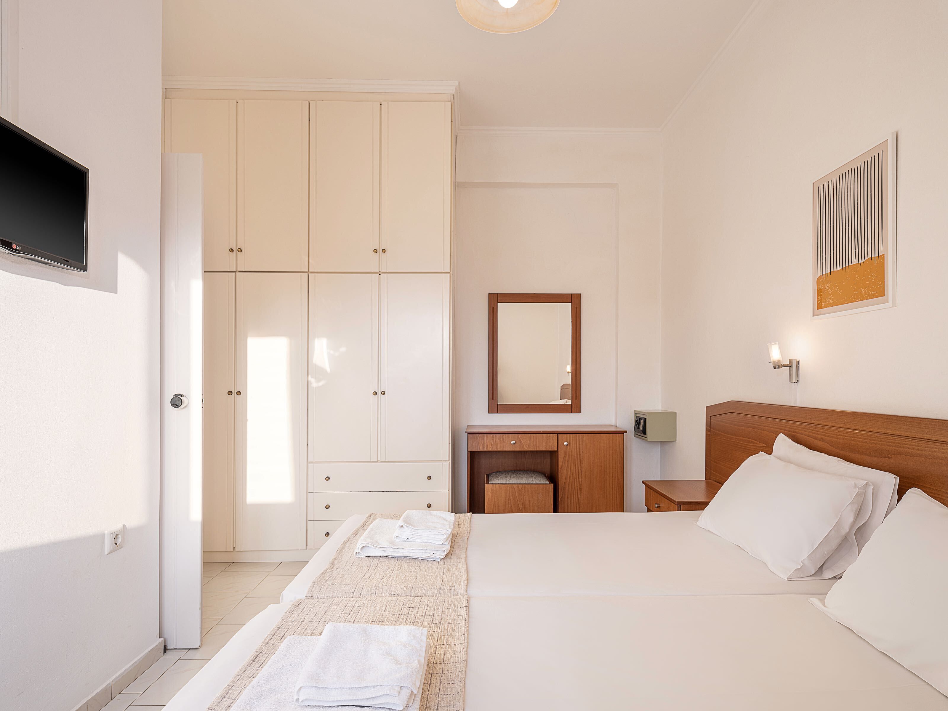 Una habitación de hotel con una cama, una mesita de noche de madera, un escritorio con una silla, un armario, un espejo y un televisor montado en la pared.