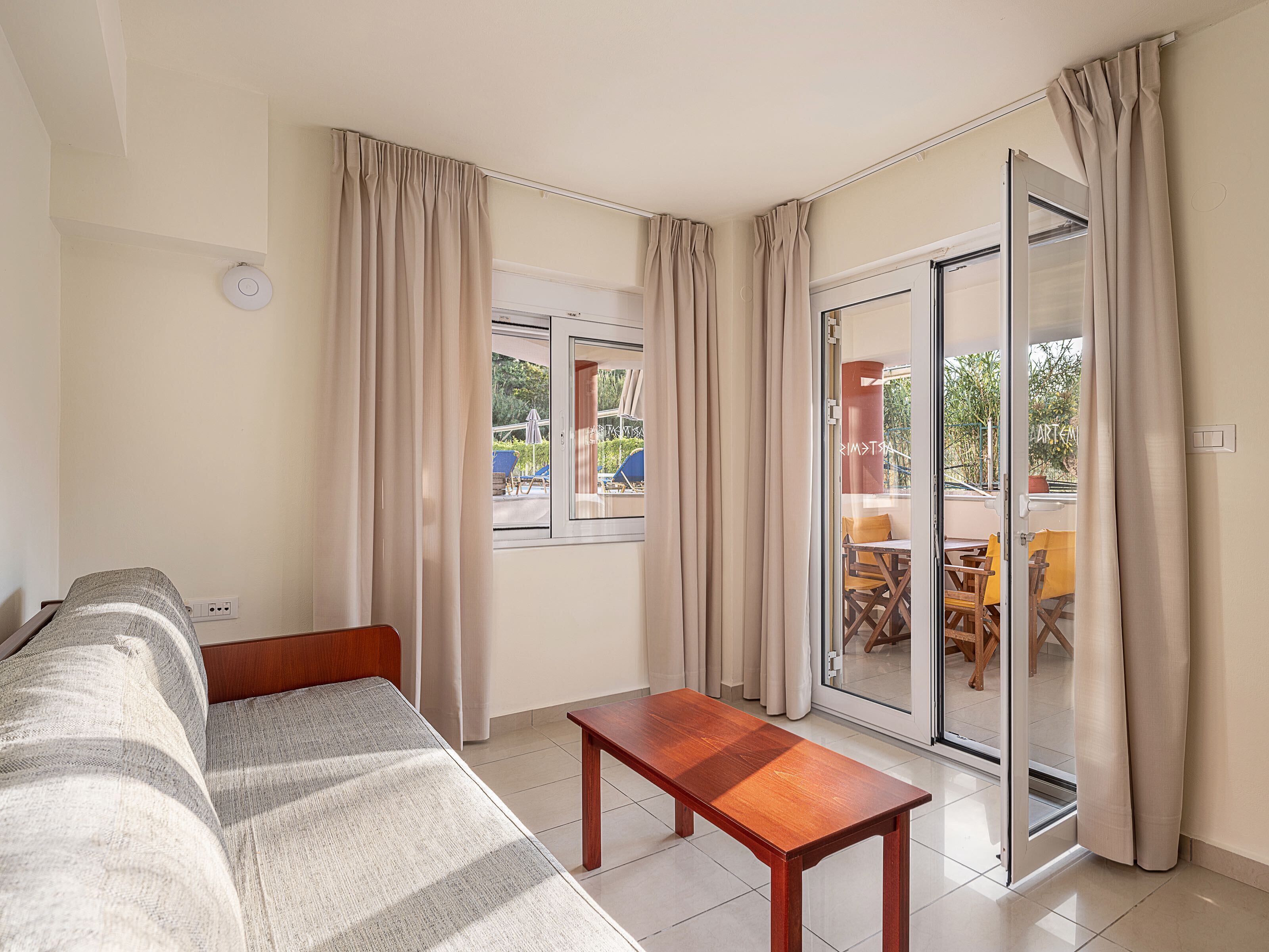 A room with a sofa, a wooden table, and a sliding glass door leading to an outdoor area with chairs and a table.