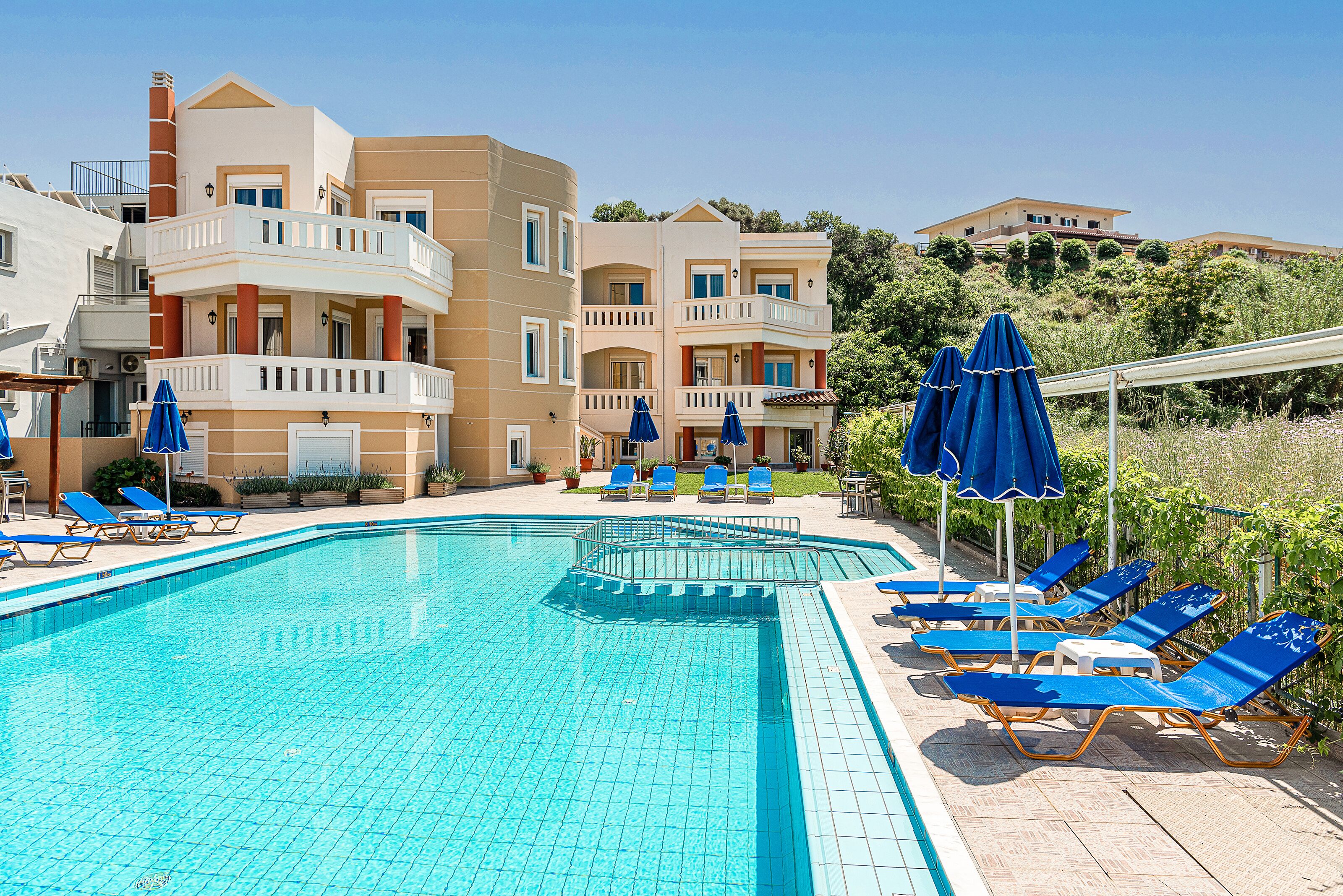 Una piscina con sillas reclinables azules y sombrillas, rodeada de edificios con balcones.
