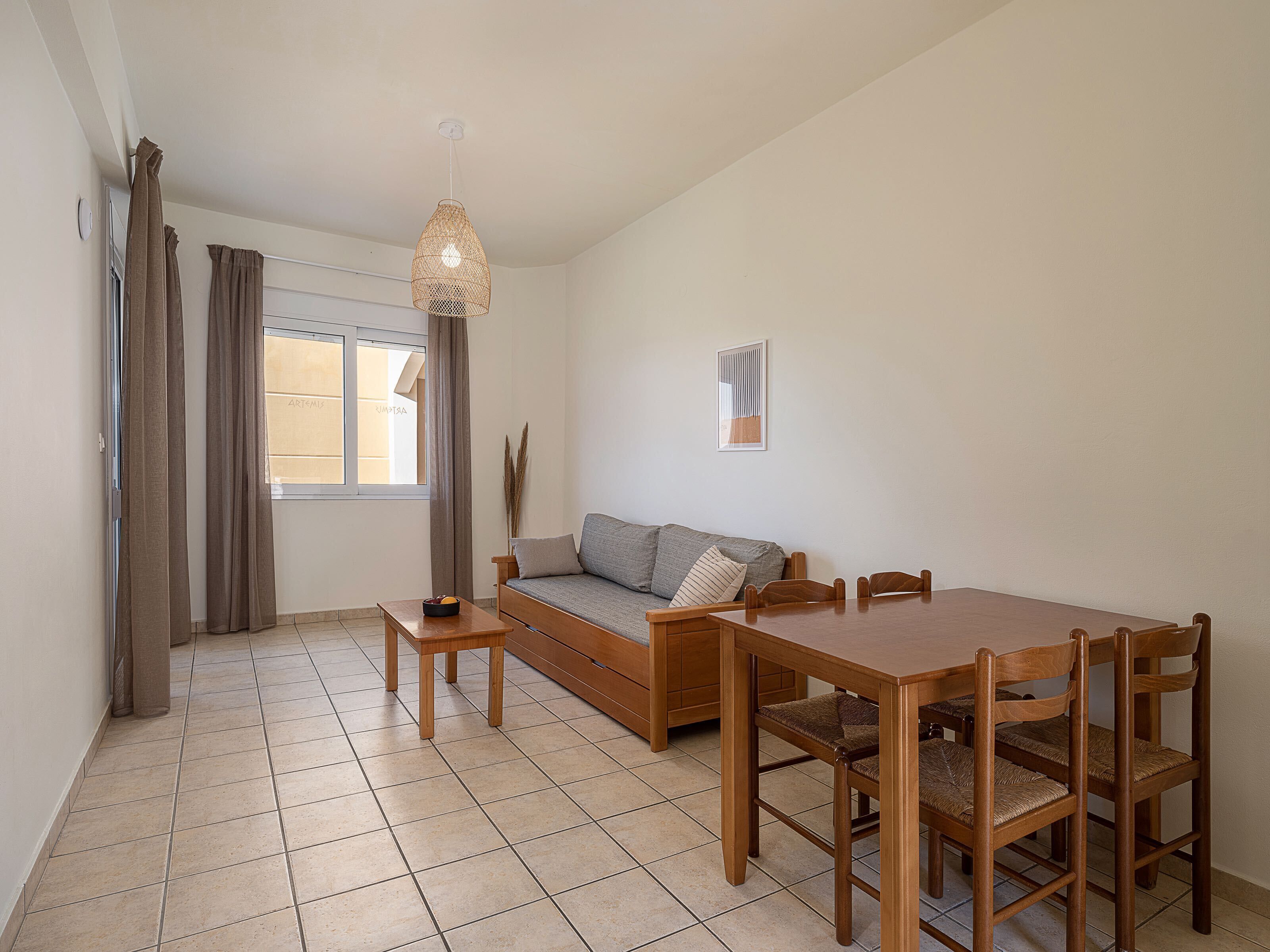 A modern living room with a sofa, dining table, and chairs. There is a window with curtains, a hanging light fixture, and a framed picture on the wall.