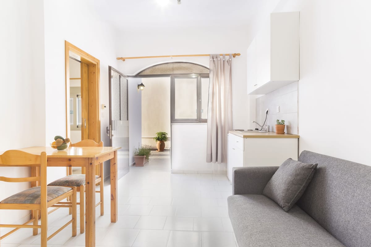 A modern, well-lit room with a dining table, chairs, a grey sofa, and a kitchen area with cabinets and a countertop.