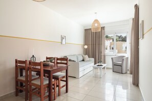A modern living room with a dining table, sofa, and armchair, a white floor, and a view through a window. - Artemis Apartments (Chania)