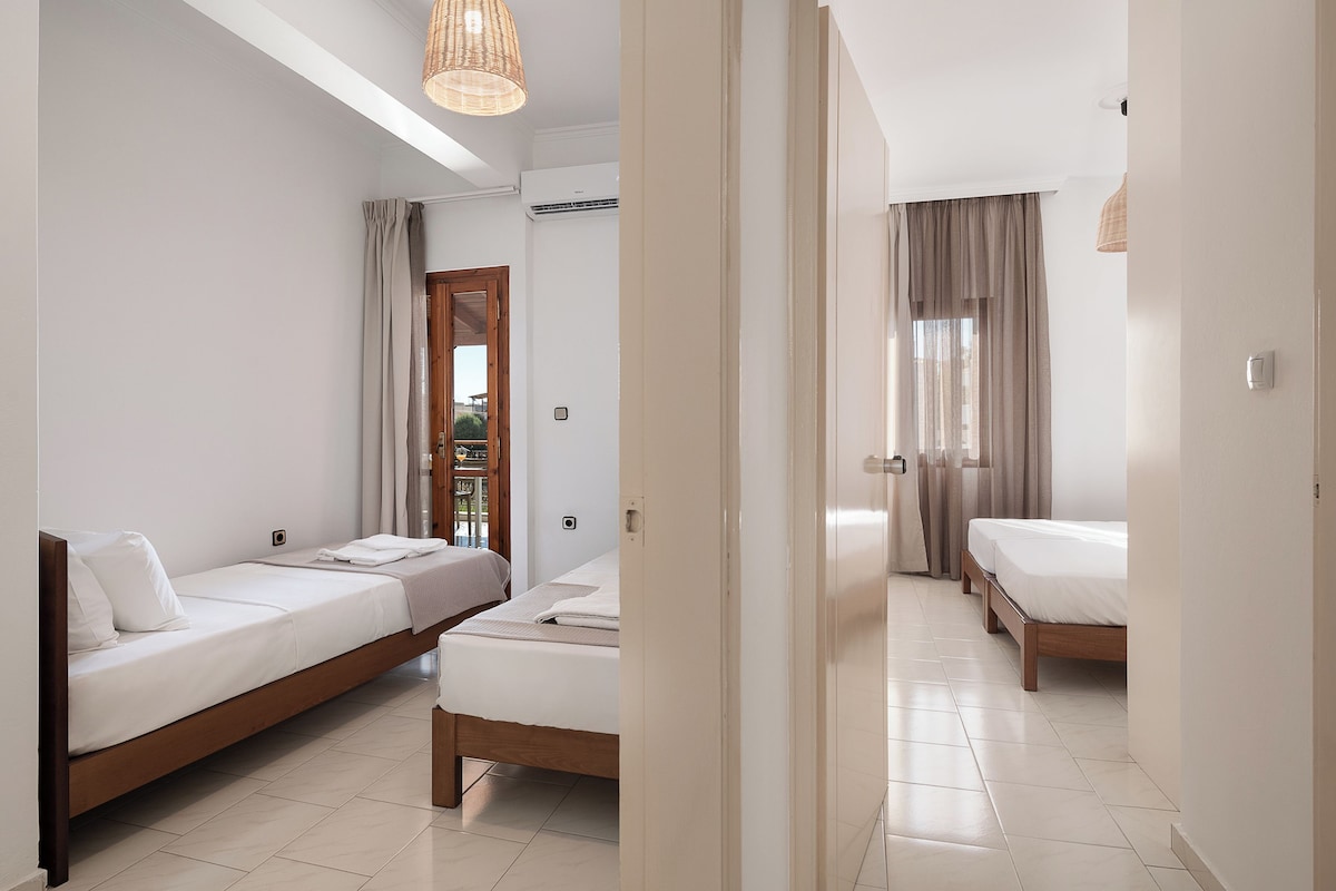 A modern hotel room with two single beds, a wooden door, and a view of the outdoors.
