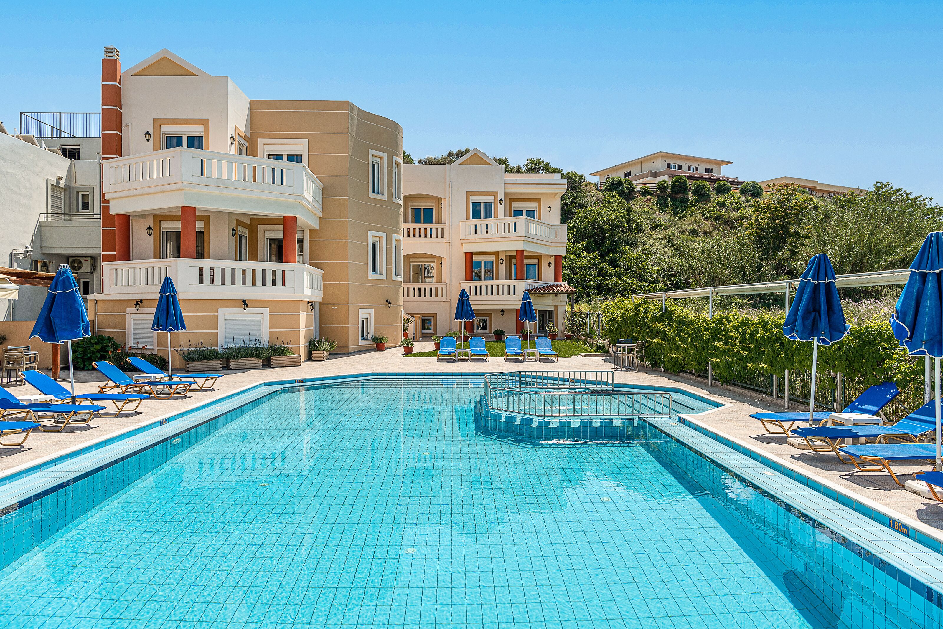 A swimming pool with blue tiles, surrounded by lounge chairs and umbrellas, and adjacent to multi-story buildings with balconies.