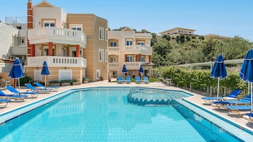A swimming pool with blue tiles, surrounded by lounge chairs and umbrellas, and adjacent to multi-story buildings with balconies.