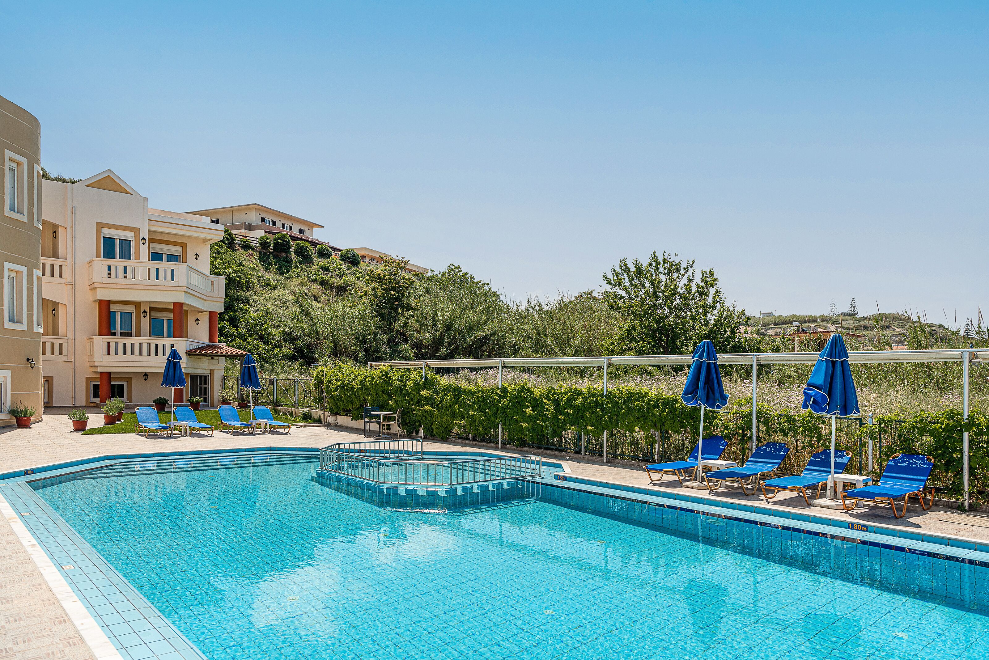 A swimming pool with blue chairs and umbrellas, surrounded by a building with a balcony and a view of a hill.