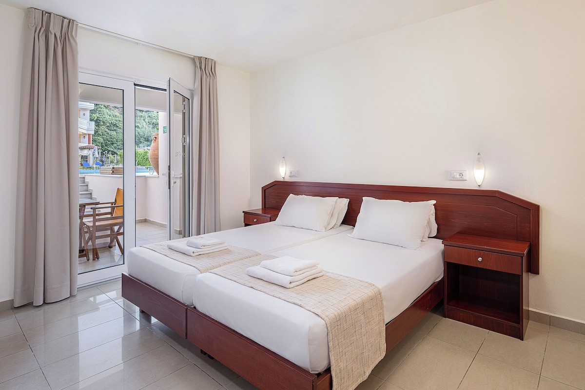 A hotel room with a large bed, two bedside tables, a wooden headboard, and a sliding glass door leading to an outdoor area with a pool and chairs.
