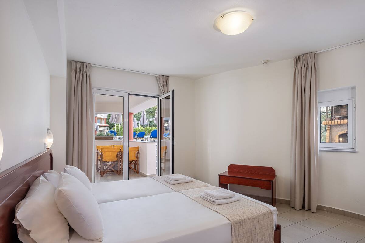 A hotel room with a bed, a wooden nightstand, a desk, and a window with curtains.