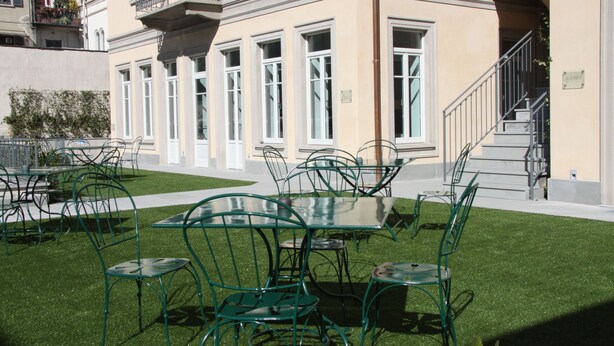 An outdoor seating area with green metal chairs and tables, a staircase, and a building with large windows and a balcony. - Hotel di Varese (Varese)